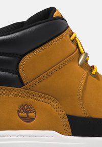 Scarpa a metà in suede marrone con accenti neri, caratterizzata da lacci gialli e rossi, occhielli in metallo e un logo ad albero marrone impresso sul lato.