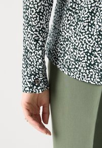 Gros plan d’une main à côté d’une manche de chemise avec un motif abstrait noir et blanc et un poignet boutonné, associé à un pantalon vert clair.
