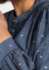 Modrý denimový top s kulatým výstřihem, zapínáním na knoflíky a nařasenými rukávy. Bílý květinový vzor dodává texturu a detail.