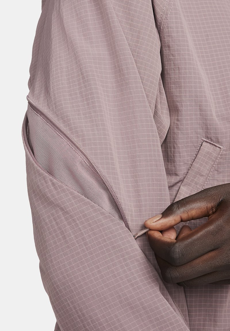 Lichtpaarse jas met een ruitpatroon en een mesh-pocketdetail op de mouw; de stof lijkt licht en textuur te hebben.