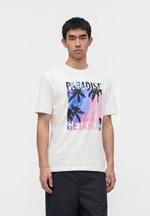 Młody mężczyzna w białej koszulce z grafiką zachodu słońca z palmą i napisem „Paradise Getaway”, w połączeniu z ciemnymi szortami.