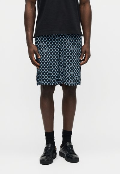 Άνδρας με μαύρο πουκάμισο, μπλε σορτς με σχέδια, μαύρες κάλτσες και μαύρα αθλητικά παπούτσια, στέκεται μπροστά σε λευκό φόντο.