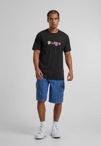 Černé bavlněné tričko s barevným grafickým potiskem "homeboy"; zkombinováno s modrými džínovými cargo šortkami s bočními kapsami; bílé tenisky.