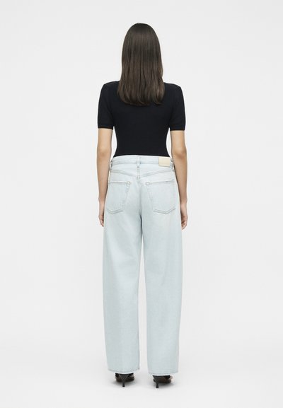 Γυναίκα με ίσια καστανά μαλλιά, φοράει μαύρο τοπ με κοντά μανίκια, ανοιχτό μπλε τζιν με φαρδιά πόδια και μαύρα σανδάλια με τακούνι, στέκεται γυρισμένη από την πλάτη.