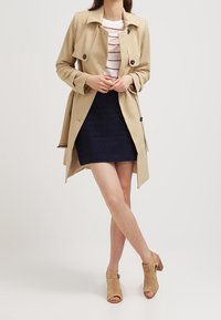 Manteau beige à gros boutons, porté sur un haut rayé blanc et rouge, associé à une jupe navy texturée et des talons ouverts couleur beige.