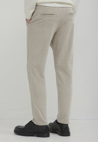 Persona con pantaloni di velluto a coste beige, maglione bianco, scarpe in pelle nera, in piedi con le mani in tasca su uno sfondo neutro.