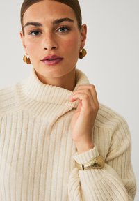 Crème kleurige geribde gebreide trui met een hoge kraag en een getextureerde gouden armband. Het model heeft een egale huid en neutrale make-up.