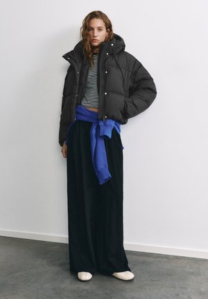 Jeune femme portant une doudoune noire, un pantalon large noir, un crop top rayé et un sweat-shirt bleu noué à la taille, debout contre un mur blanc.