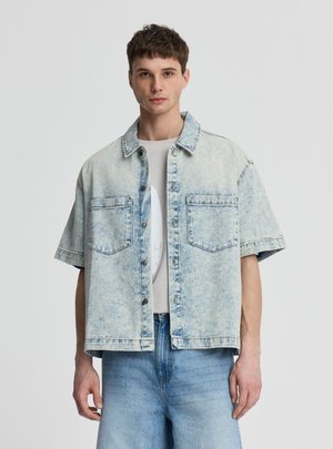 Giovane uomo che indossa una giacca di jeans oversize a maniche corte in lavaggio chiaro sopra una t-shirt bianca con grafica e jeans azzurri, in piedi davanti a uno sfondo semplice.