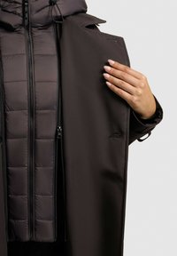 Una mano che sistema il risvolto di un cappotto marrone scuro indossato sopra una giacca nera trapuntata con cerniere visibili su uno sfondo grigio uniforme.
