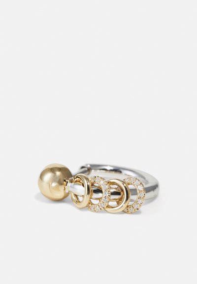 Guld- och silverring med tre sammanlänkande gyllene cirklar, där en är prydd med klara stenar. Har en slät, polerad yta och en sfärisk detalj.
