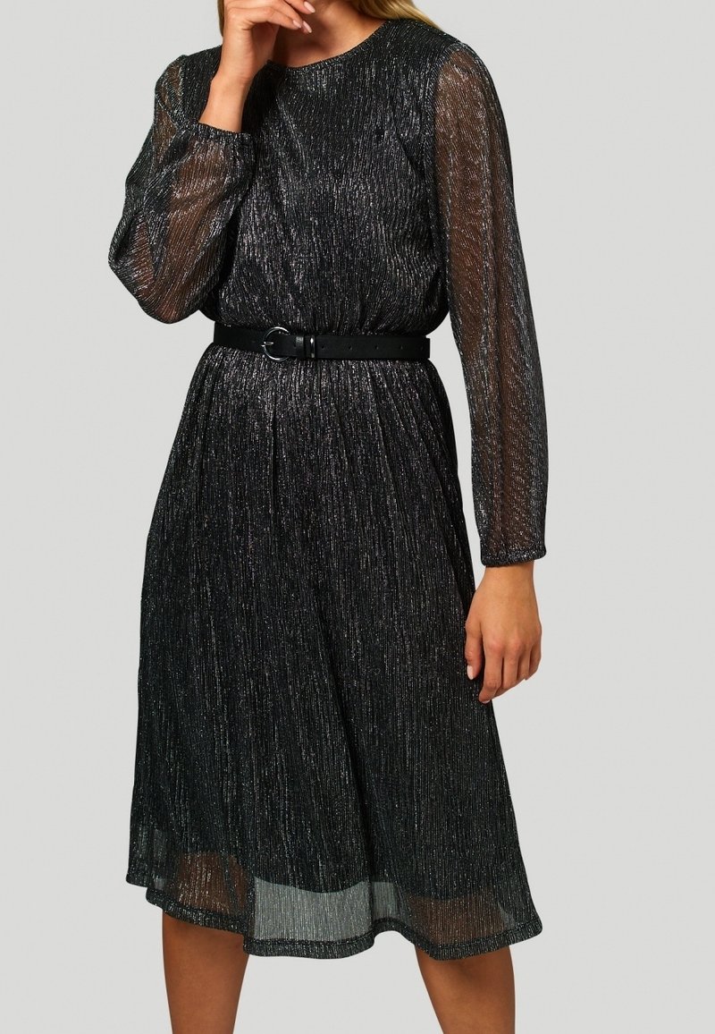 Vestido negro con mangas largas transparentes, tejido texturizado con hilos plateados, cintura ceñida con un cinturón negro, falda a la altura de la rodilla.