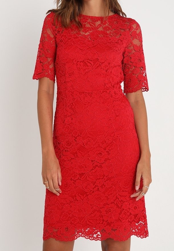 Femme portant une robe en dentelle rouge mi-longue à manches courtes avec des motifs floraux, debout devant un fond clair uni.