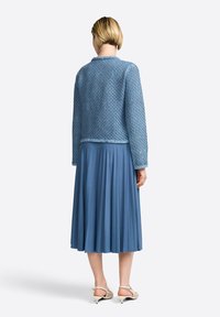 Giacca a maglia azzurro chiaro con un motivo testurizzato, abbinata a una gonna blu plissettata. La modella indossa sandali con tacco beige.