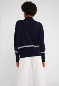 Person mit lockigem Haar, die goldene Creolen, einen dunkelblauen Pullover mit grauen Streifen und weiße Hosen trägt, von hinten vor einem einfarbigen Hintergrund.
