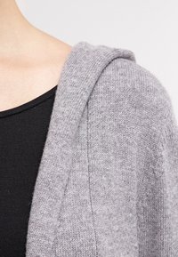 Cardigan gris de punto con textura suave y capucha. Presenta un ribete acanalado a lo largo del frente abierto, sobre una camiseta negra.