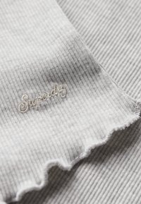 Tejido gris acanalado con un borde deshilachado, que presenta texto bordado en beige claro "Superdry" en una tipografía cursiva. Textura suave, diseño informal.