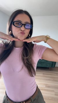 Mustat ylisuuret lasit sinisellä sävyllä, vaaleanpunainen tiukka t-paita, kultaiset renkaat, ja kultakello; kädet alaspäin leuan alla leikkisässä asennossa.