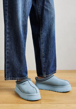 Kék velúr slip-on UGG cipők vastag talppal, kék farmerrel viselve, fából készült padlón, sima háttér előtt.