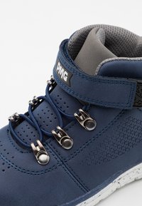 Botte montante bleu marine avec tige en cuir, col rembourré gris et accessoires métalliques pour les lacets. Détail latéral texturé et semelle mouchetée.