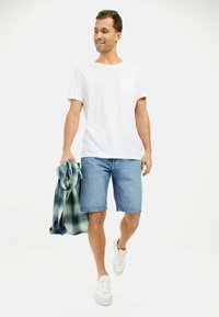 Hvid kortærmet T-shirt med lomme, blå denim shorts og hvide sneakers. Holder en grønternet skjorte. Neutral baggrund.