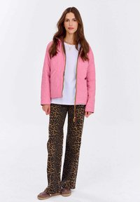 Różowa pikowana kurtka z złotym zamkiem, w zestawieniu z szerokimi spodniami w print lamparta. Modelka stoi na jednolitym tle, nosząc brązowe buty.