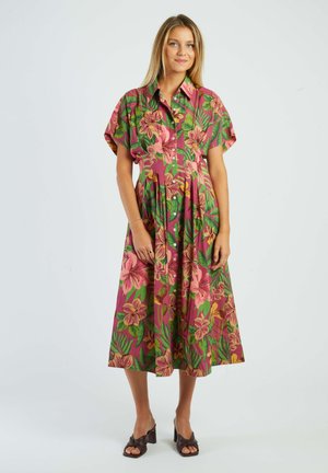 Frau steht in einem knielangen, kurzärmeligen Kleid mit pink-grünem Blumenmuster, Knopfleiste vorne und braunen Sandaletten mit Absatz.