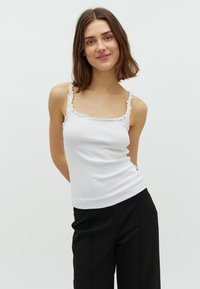 Witte geribbelde tanktop met rimpel-laceafwerking langs de halslijn en schouderbanden, gedragen met zwarte losse broeken. Glad textuur en slanke pasvorm.