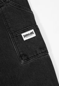 Juodas džinsinis audinys turi matomą siuvimą, šoninį kišenę su stačiakampiu baltu etikečių, kuris skaito "wasted" ir tekstu "ALWAYS FAITHFUL."