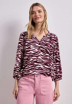 Kobieta w różowo-czarnej bluzce w paski zebry z długim rękawem i dekoltem w serek oraz różowych spodniach, stojąca na neutralnym tle.