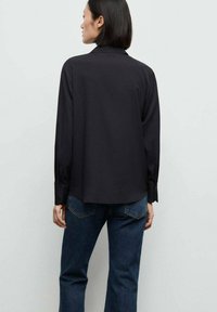Svart långärmad skjorta med krage, enkel design och slät textur. Bärs med mörkblå jeans som visar en avslappnad passform och knäppta manschetter.