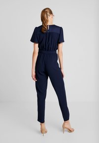 Mujer con coleta, vestida con un mono azul marino y sandalias de tacón nude, de pie frente a un fondo gris claro liso.