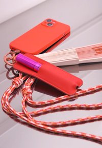 Funda de teléfono de silicona roja junto a un bolígrafo rosa en un estuche de cuero rojo, con un cordón trenzado multicolor, todo sobre una superficie reflectante.