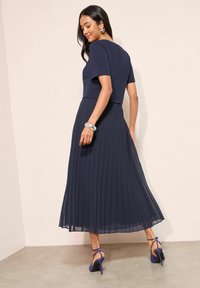 Sötétkék ruha, testhezálló felsőrésszel és redős szoknyával, könnyű anyagból készült. Lakásban lila színű, bokapántos magas sarkú cipőkkel párosítva.