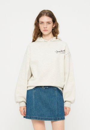 Kobieta w kremowej bluzie z kapturem z napisem "Sweet Good" i nitowanymi detalami, połączona z niebieską spódnicą jeansową z czarnym paskiem, stojąca na białym tle.