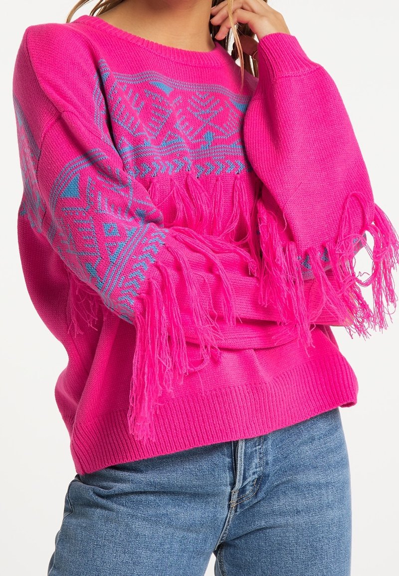 Pull rose vif avec motifs géométriques bleus et longues franges aux manches, porté avec un jean bleu, debout avec une main près du visage.