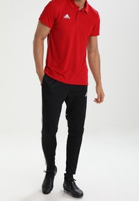 Rotes Poloshirt mit Kragen und drei Knöpfen, kombiniert mit schwarzen Sporthosen mit weißen Seitenstreifen und schwarzen Fußballschuhen.