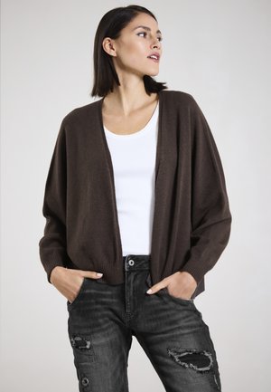 Braune, offen geschnittene Strickjacke aus weichem Material mit gerippten Bündchen, kombiniert mit einem weißen Tanktop und ausgebeulten schwarzen Jeans.
