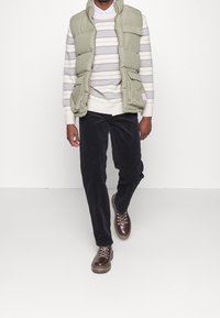 Vadderad olivgrön väst över en randig grå och vit tröja, i kombination med mörka kordbyxor och bruna snörstövlar. Enkelt mönster och textur.