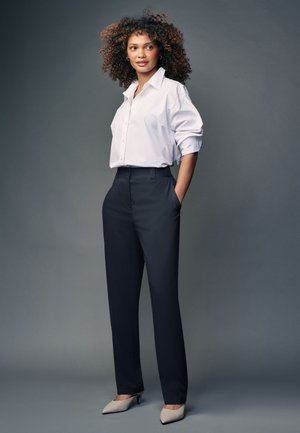 Mujer con cabello rizado que lleva una camisa blanca abotonada, pantalones negros y tacones beige, de pie con las manos en los bolsillos frente a un fondo gris.