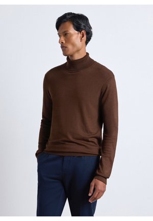 Uomo che indossa un maglione marrone a collo alto e pantaloni blu scuro, in piedi con le mani nelle tasche, guarda di lato su uno sfondo semplice.