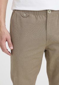 Pantalones beige con estampado y textura ligera, que cuentan con una cinturilla con botones y bolsillos laterales, mostrados a la altura de la cadera.