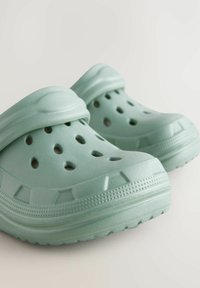 Sabots vert clair en plastique moulé, dotés de trous de ventilation, d'une surface texturée et d'une sangle de talon sécurisée pour plus de stabilité.