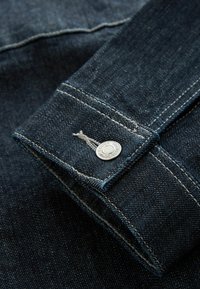 Donkerdenimstof met contrasterende stiksels, een knoop met gravure en een opgerolde manchet. De textuur ziet er glad uit met subtiele kleurnuances.