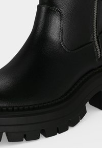 Gros plan d'une bottine noire en cuir avec une semelle épaisse crantée et une fermeture éclair latérale sur un fond clair.