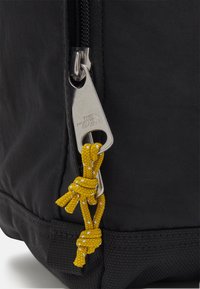 Gros plan sur la fermeture éclair d’un sac à dos noir avec une tirette argentée gravée "The North Face" et un cordon jaune noué attaché.