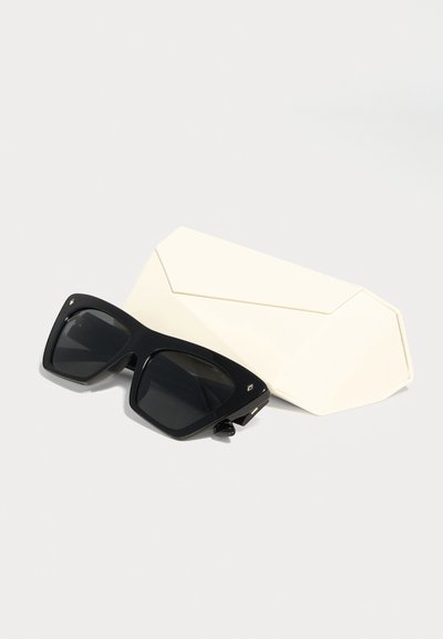 Μαύρα υπερμεγέθη ορθογώνια γυαλιά ηλίου δίπλα σε μία λευκή γεωμετρική σκληρή θήκη πάνω σε φωτεινό φόντο.