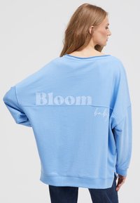 Light blue oversized sweatshirt with a wide neckline, droopy shoulders, and "Bloom" and "baby" printed in white on the back. Soft fabric texture.