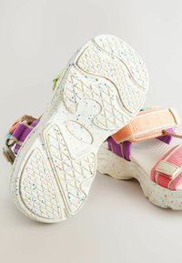 Farebné sandále majú textúrovanú bielu podrážku s multicolornými škvrnami. Nastaviteľné popruhy v ružovej, fialovej a zelenej farbe zabezpečujú bezpečné uchytenie.