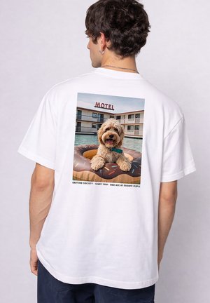 Mann trägt weißes T-Shirt mit Foto eines Hundes auf einem Donut-Schwimmring im Pool vor einem Motel, Rückansicht.